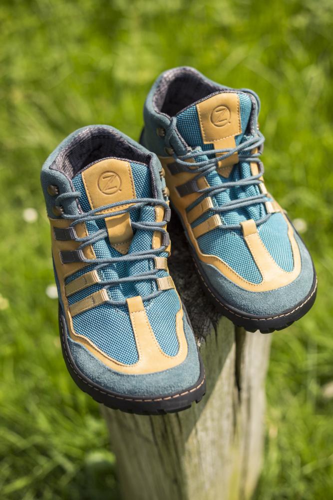 Ein Paar ESQAPE Blue Orange Waterproof Wanderschuhe von ZAQQ, mit einem auffälligen blau-beigen Farbschema mit schwarzen Sohlen und dem unverwechselbaren ZAQQ TERRA Sohlendesign, ruht auf einem Holzpfosten. Diese wasserdichten Schuhe heben sich vor einem Hintergrund aus grünem Gras ab und vereinen Stil und Funktionalität mit ihren mehreren Riemen für jedes Abenteuer.