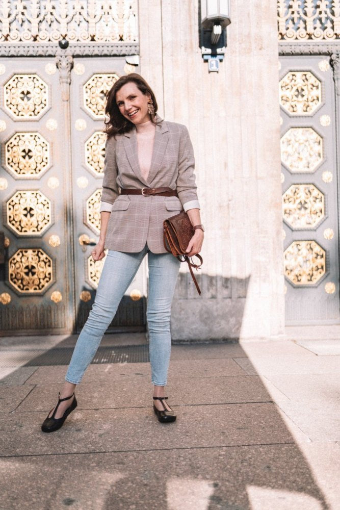 Eine Frau in kariertem Blazer, Jeans und schwarzen Schuhen mit Barfußsohle steht selbstbewusst vor einer verzierten Tür mit sechseckigem Muster. Sie lächelt und hält eine dunkelbraune QITE-Clutch von ZAQQ, während das Sonnenlicht Schatten auf den Boden wirft.