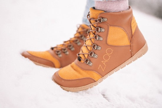 Auf einer schneebedeckten Fläche ist ein Paar braun-orangefarbene Stiefel mit der Aufschrift ZAQQ zu sehen. Die Stiefel haben beigefarbene Sohlen und sind geschnürt. Der Träger ist teilweise zu sehen; er trägt hellblaue Jeans. Die Szene wirkt hell und winterlich.