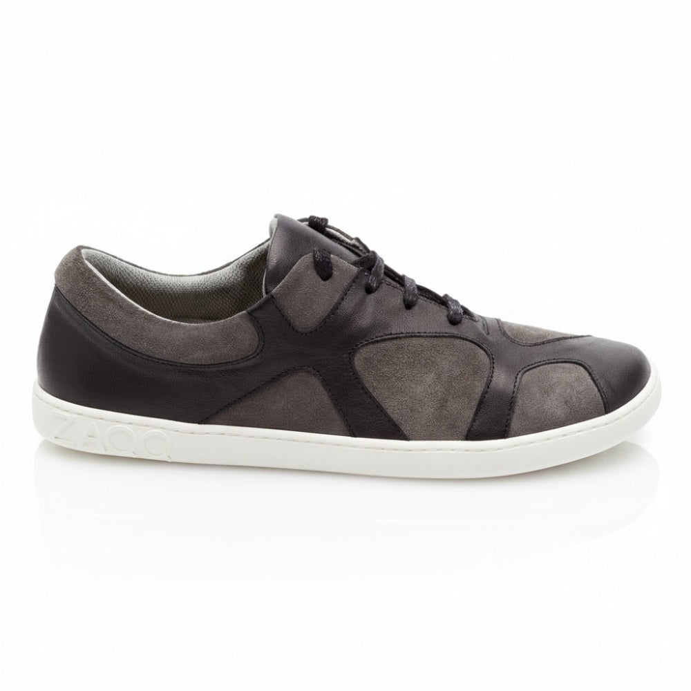 Ein einzelner Sneaker in Dunkelgrau und Schwarz aus der QAAANAAQ Black-Kollektion von ZAQQ weist ein strukturiertes Design auf dem Obermaterial auf und hat den Markennamen seitlich auf der Sohle eingeprägt. Er ist aus Veloursleder für einen leichten, abgenutzten Look gefertigt und weist flache Schnürsenkel und einen Low-Cut-Stil auf, der sich vor einem weißen Hintergrund abhebt.
