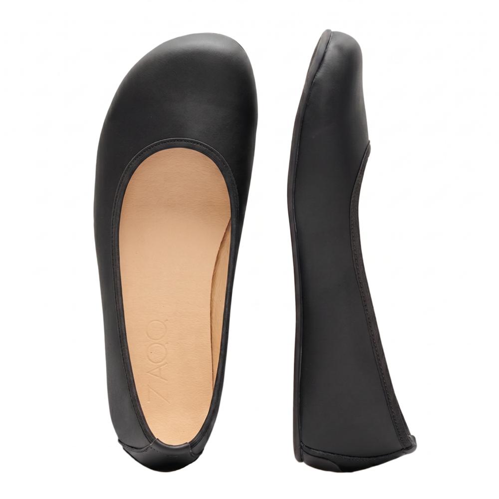 Ausgestellt wird ein Paar schwarze Ballerinas ZAQQ QARMA. Ein Schuh liegt auf der Seite und zeigt die glatte schwarze Sohle und das Fußbett, während der andere aufrecht steht und eine glatte Lederoberfläche offenbart, die an die Anmut einer Ballerina erinnert, ergänzt durch ein warmes braunes Innenfutter.