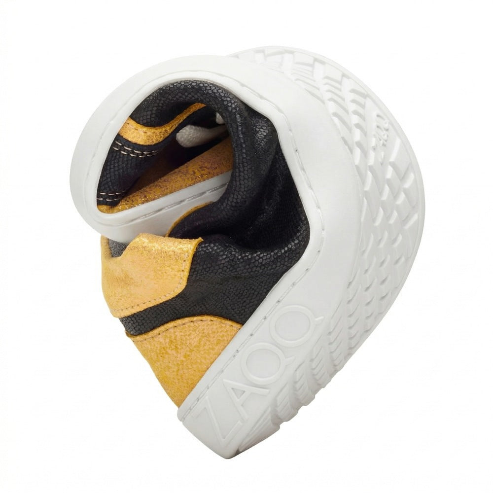 Der Barfußschuh ist ein herzförmiger, kunstvoll gefalteter Schuh, der seine Flexibilität und seinen außergewöhnlichen Komfort demonstriert. Er ist aus schwarzem Material mit goldenen Akzenten gestaltet. Er verfügt über eine strukturierte weiße Sohle mit dem geprägten Markennamen ZAQQ und ist als QODY Gold bekannt.